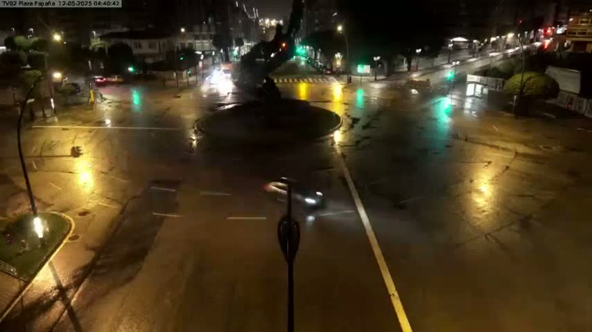 A nighttime, rain-soaked intersection shows several cars navigating a flooded roundabout and surrounding streets, illuminated by streetlights.
