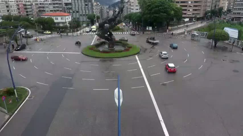 A wet roundabout with several cars navigating it, centered around a large, dark-colored statue.