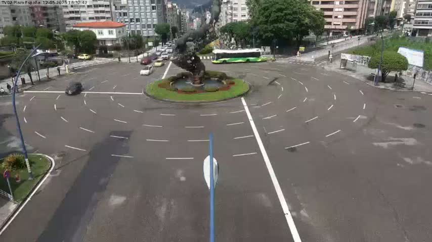 A traffic circle with a large sculpture in the center is surrounded by buildings and a few cars, under a partly cloudy sky.