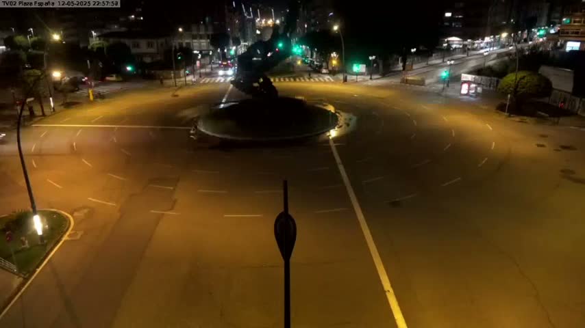 A mostly empty roundabout at night, illuminated by streetlights, features a dark statue in its center.