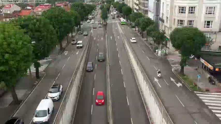 A wet, multi-lane road with moderate traffic, bordered by green trees and buildings.
