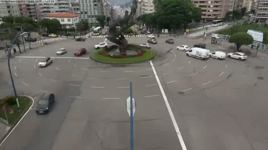 A roundabout with several cars navigating it, surrounded by buildings and trees under an overcast sky.