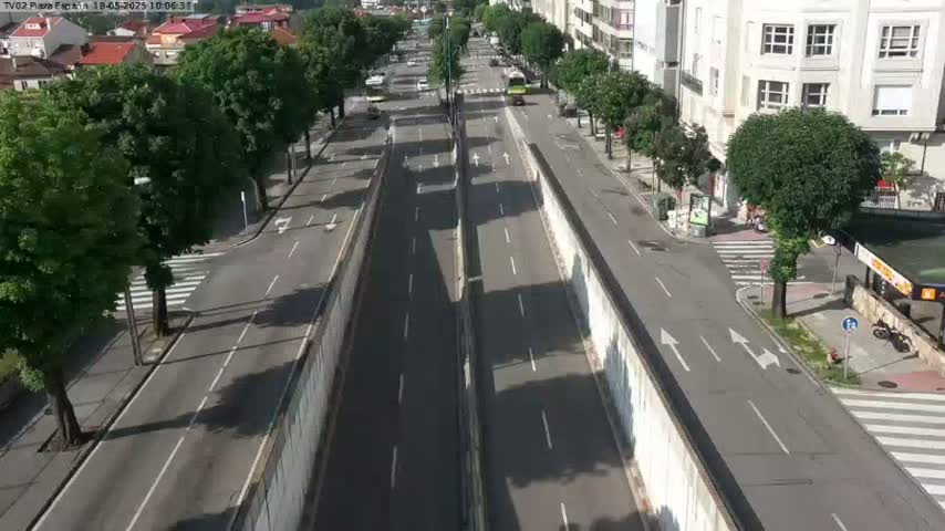 A mostly empty, two-lane road lined with leafy green trees runs through a city under a sunny sky.