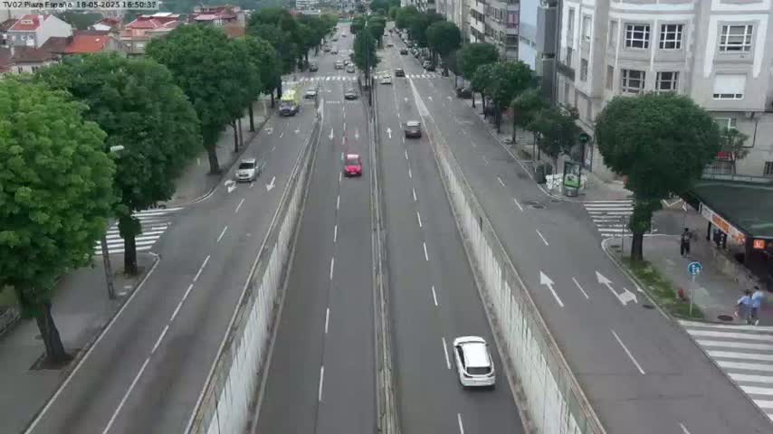A two-lane road with several cars driving on it, flanked by trees and buildings on either side, on a seemingly clear day.