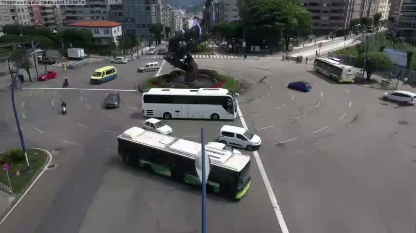 A roundabout with several cars, buses, and motorbikes navigating it under a mostly sunny sky.