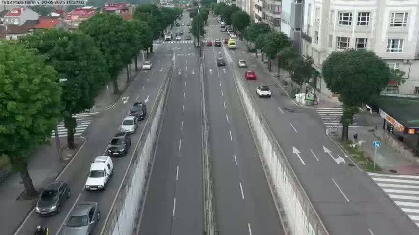 A multi-lane road with light traffic is seen from an elevated perspective, flanked by green trees and buildings, under what appears to be an overcast sky.