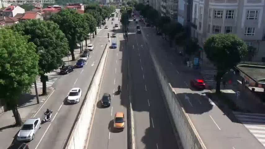 A sunny day shows a multi-lane road with cars and a motorcycle driving in both directions, flanked by a tree-lined street and tall buildings.