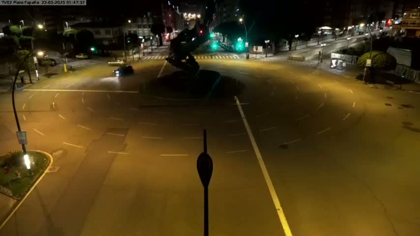 A nighttime aerial view shows a mostly empty roundabout with a dark sculpture in the center, surrounded by roadways and buildings.