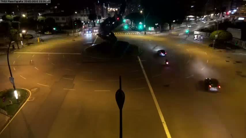 A nighttime, high-angle view of a roundabout with several vehicles and a few pedestrians navigating it under slightly hazy conditions.