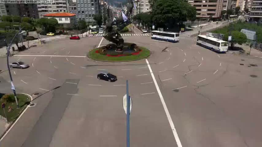 A high-angle view shows a roundabout with several cars and buses, under sunny conditions.