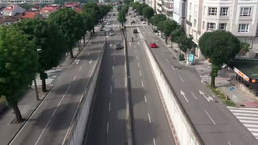 A mostly sunny, high-angle view of a multi-lane road with sparse traffic, flanked by sidewalks lined with trees and buildings.