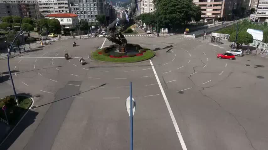 A mostly empty roundabout with a large sculpture in the center, a few vehicles, and pedestrians, on a sunny day.