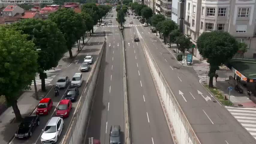 A high-angle view of a multi-lane road with moderate traffic on a sunny day, flanked by buildings and lined with trees.