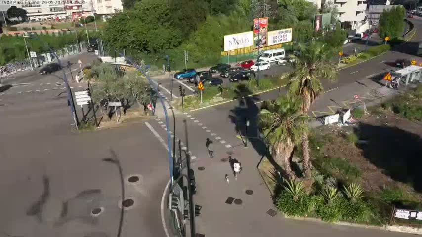 An aerial view of a sunny intersection with several cars parked and driving, pedestrians walking, and palm trees in landscaped areas.