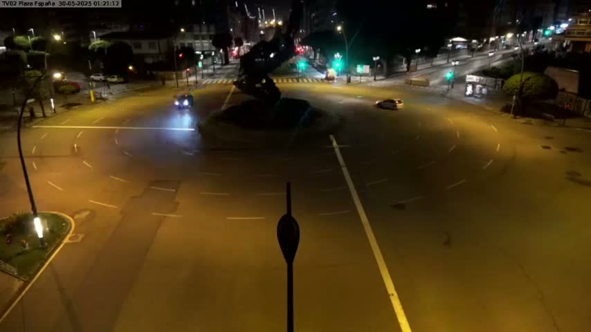 At night, a few cars are driving around a roundabout in front of a large dark statue.