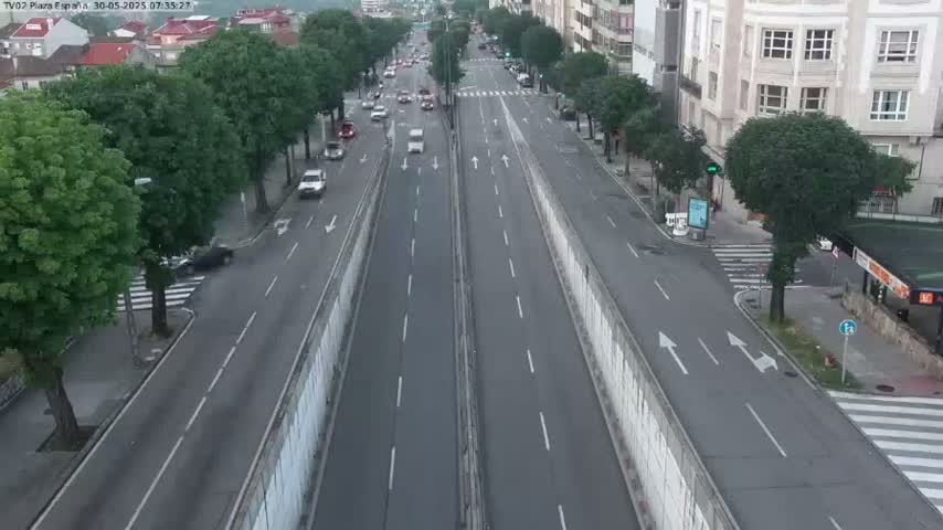 A high-angle view shows a multi-lane road with sparse traffic, flanked by buildings and leafy green trees under a seemingly clear sky.