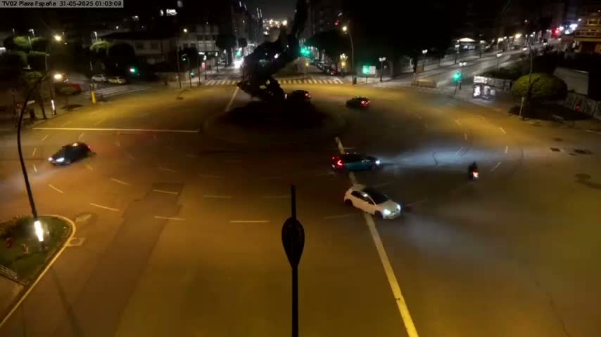 A high-angle nighttime view shows several cars navigating a roundabout, with one car appearing to be drifting, and a motorcycle also present.