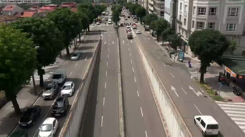 A sunny day reveals a view of a multi-lane road with moderate traffic, lined with buildings and trees.
