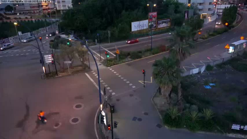 An aerial view of a city intersection at dusk, showing cars, pedestrians, and traffic lights, with some palm trees and greenery along the roadside.