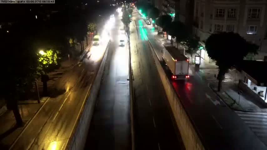 A rain-slicked, two-lane road at night is sparsely populated with vehicles, with city buildings and trees lining the sides.