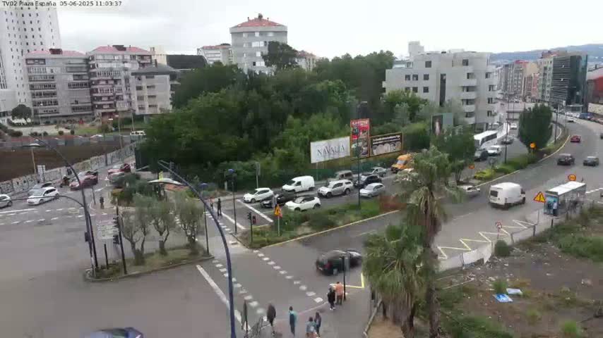 An overcast day shows a busy city intersection with multiple cars and pedestrians, surrounded by buildings and lush green vegetation.