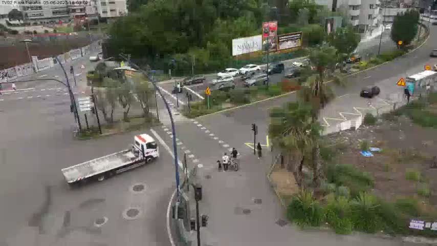 A flatbed tow truck is parked near a multi-lane intersection with several cars and pedestrians, under an overcast sky.