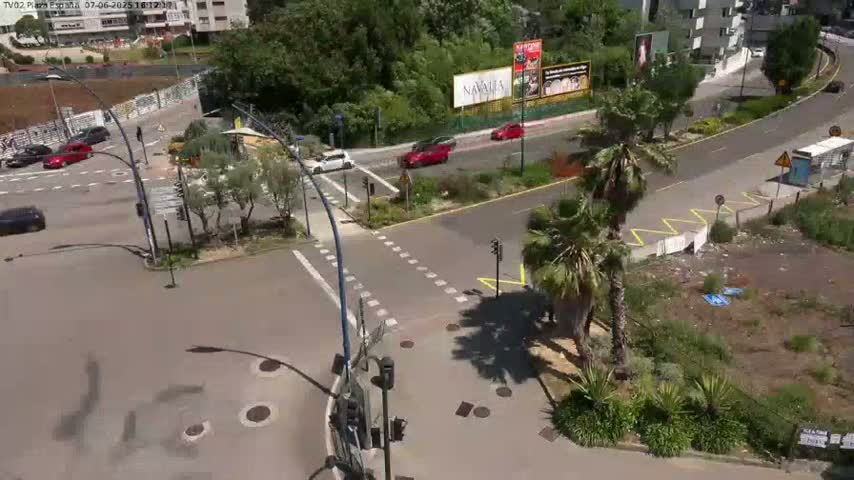 A sunny day overlooks a street intersection with several cars, surrounded by palm trees, buildings, and a vacant lot.