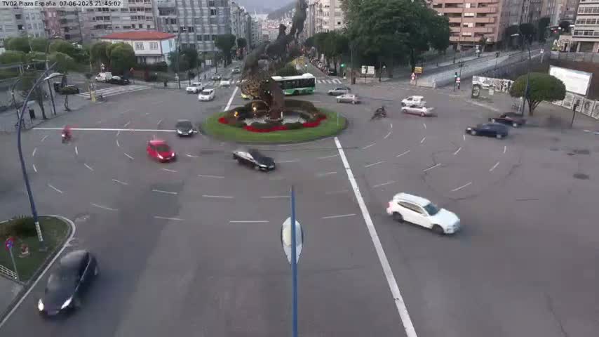 Several cars navigate a roundabout at dusk, with a large sculpture in the center.