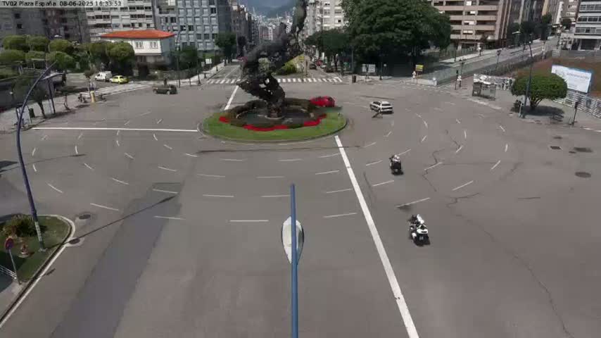 A high-angle view shows a mostly empty traffic circle with a large abstract sculpture in the center, several cars and motorcycles, and buildings in the background on a sunny day.