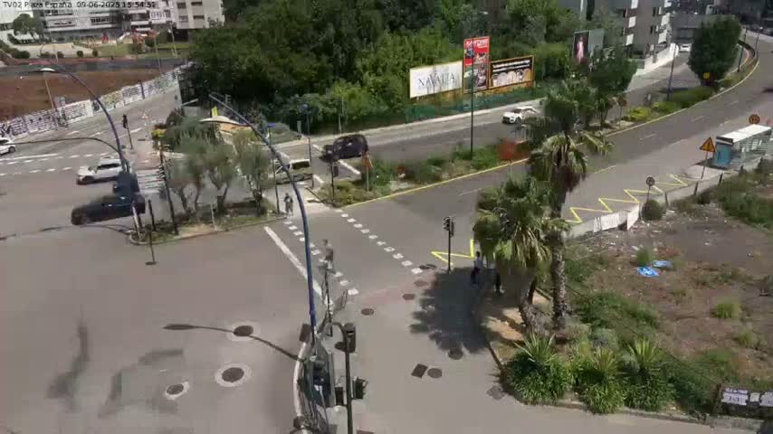 A sunny day shows a high-angle view of a multi-lane roadway intersection with several cars and pedestrians, surrounded by lush greenery and buildings.