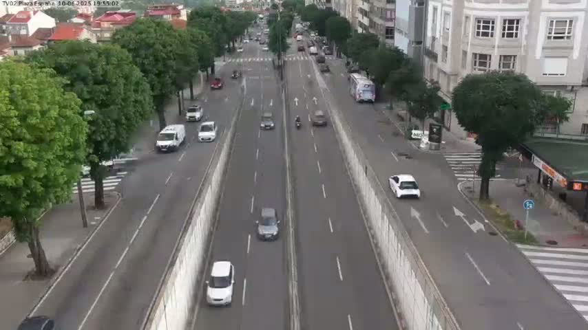 A high-angle view shows a city street with several cars driving in both directions, lined with trees and buildings on either side, under a seemingly clear sky.