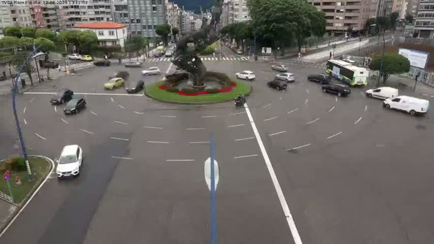 Several cars navigate a roundabout surrounding a sculpture, under overcast skies.