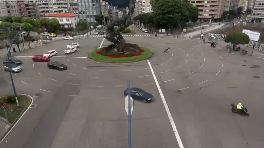 A high-angle view of a roundabout with several cars navigating it, a statue in the center, and a person on a three-wheeled vehicle in the lower right corner under sunny skies.