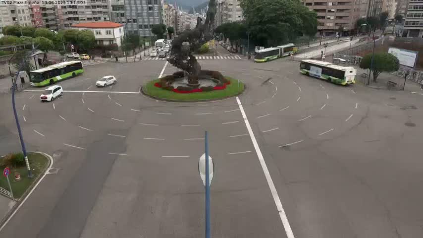 A roundabout with several cars and buses, a central sculpture, and surrounding buildings on a sunny day.