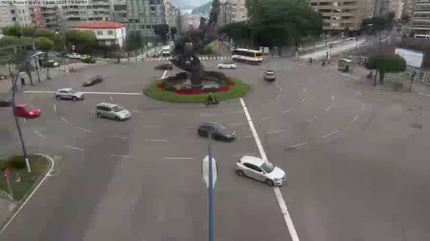 Several cars are navigating a roundabout centered on a large, dark statue, under an overcast sky.