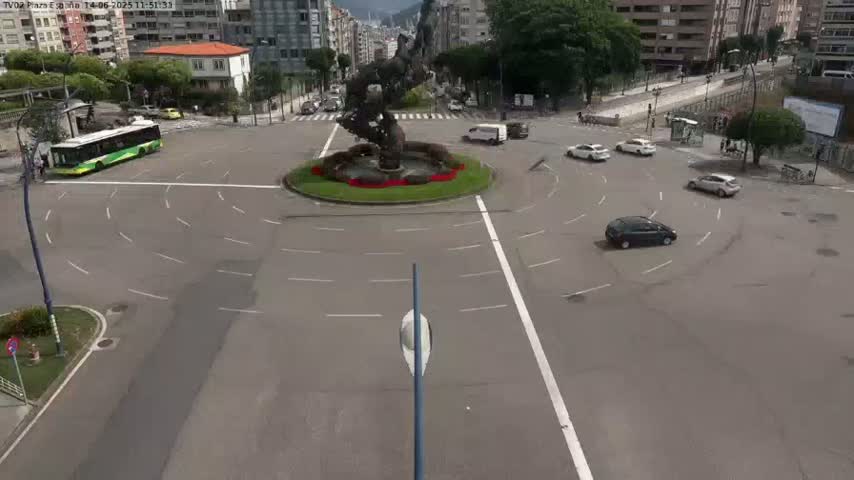 A roundabout with a large, abstract sculpture in the center, surrounded by several cars and a bus, on a mostly sunny day.