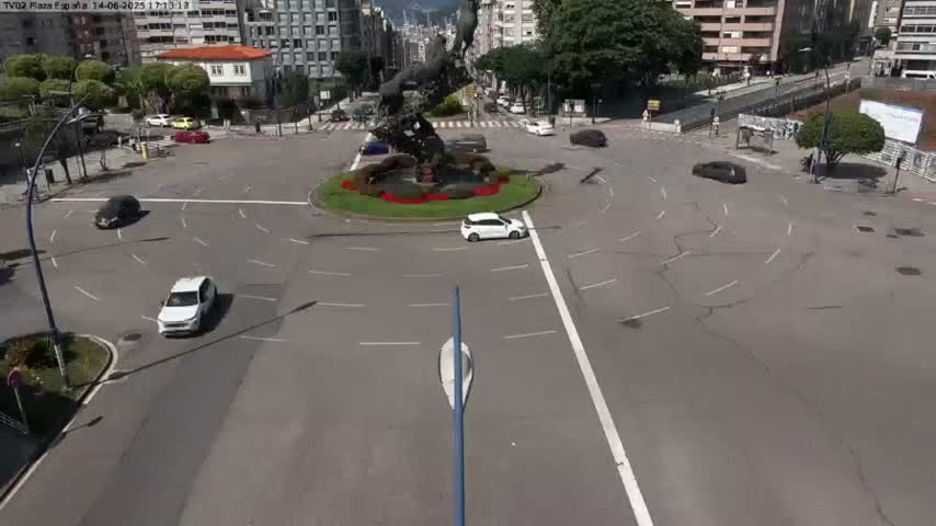 A roundabout with several cars, a central statue, and surrounding buildings on a sunny day.