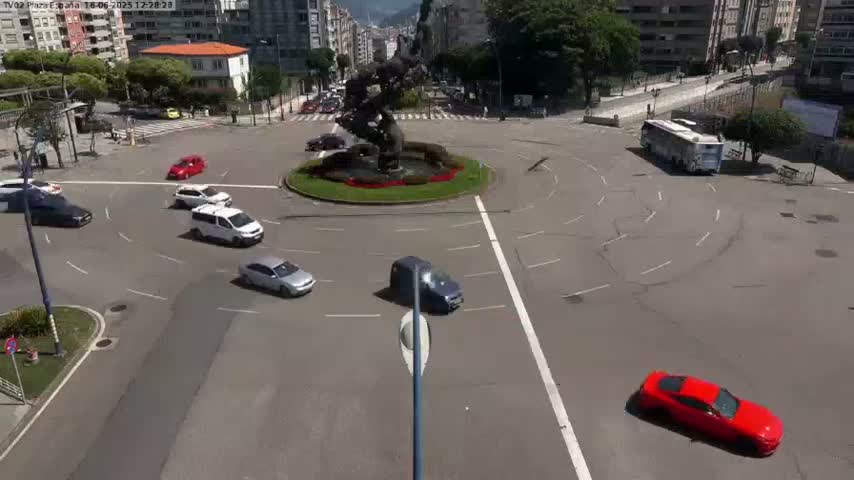 A roundabout with several cars navigating it under sunny conditions, centered around a sculpture.