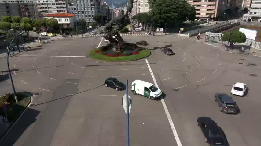 A sunny day shows several cars navigating a roundabout centered on a large, dark statue; buildings surround the roundabout.