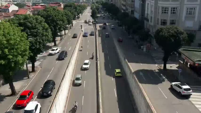 A sunny day on a city street shows several cars driving in both directions along a divided roadway lined with trees and buildings.