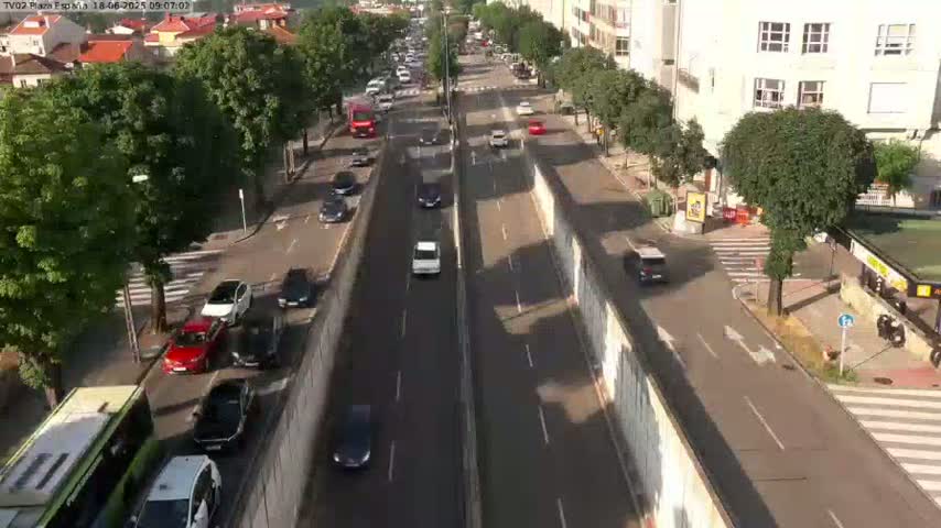 A sunny day reveals a highway scene with numerous vehicles traveling in both directions, flanked by buildings and lush green trees.