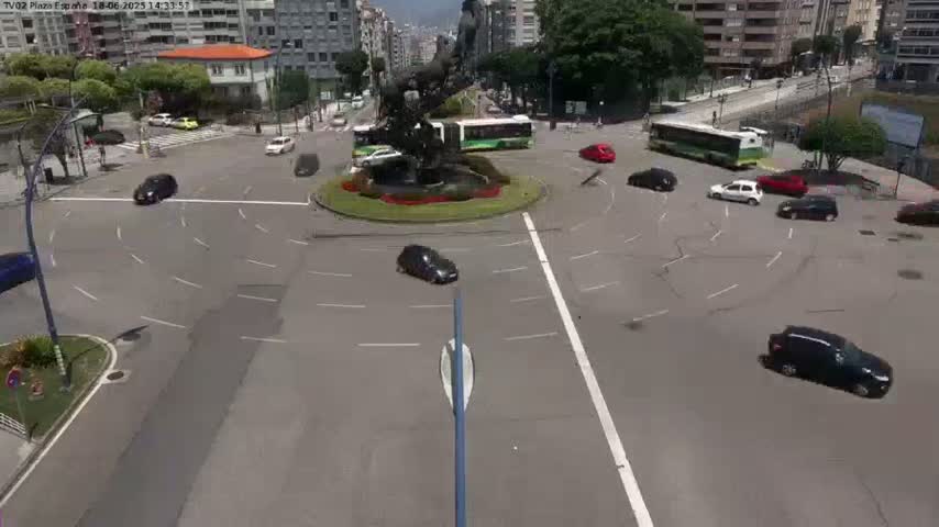 Several cars navigate a roundabout in a city, with buses visible in the background under a sunny sky.