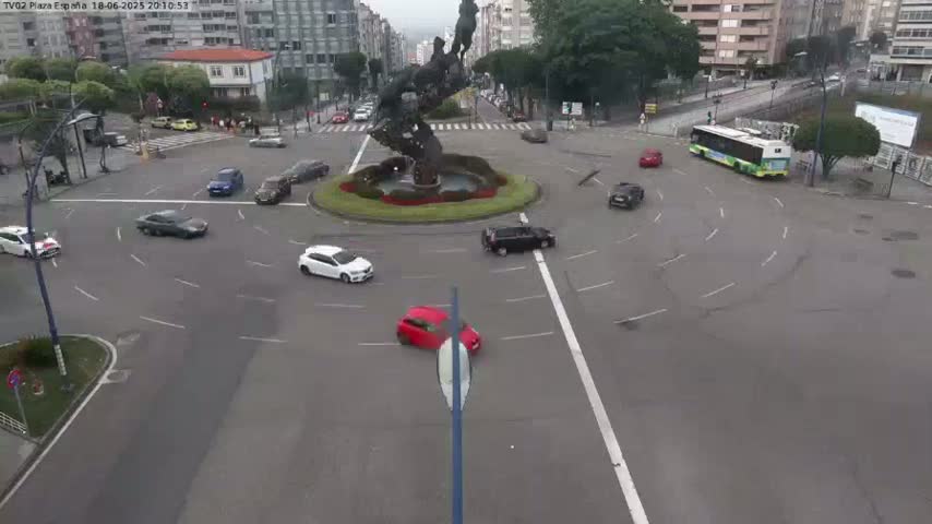 Several cars are navigating a roundabout centered on a large, abstract sculpture, under an overcast sky.
