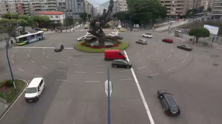 A roundabout with several cars and a bus, featuring a large metal sculpture in the center, under an overcast sky.