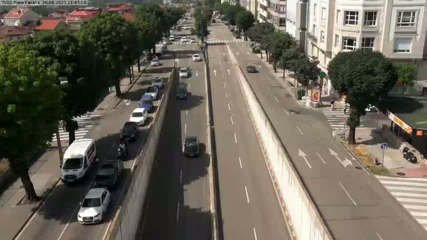 A sunny day reveals a multi-lane road, bordered by buildings and trees, with numerous cars parked and driving along it.