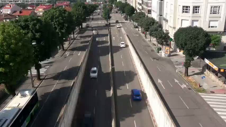 A high-angle view of a city street on a sunny day, showing several cars driving in both directions on a divided roadway lined with trees and buildings.