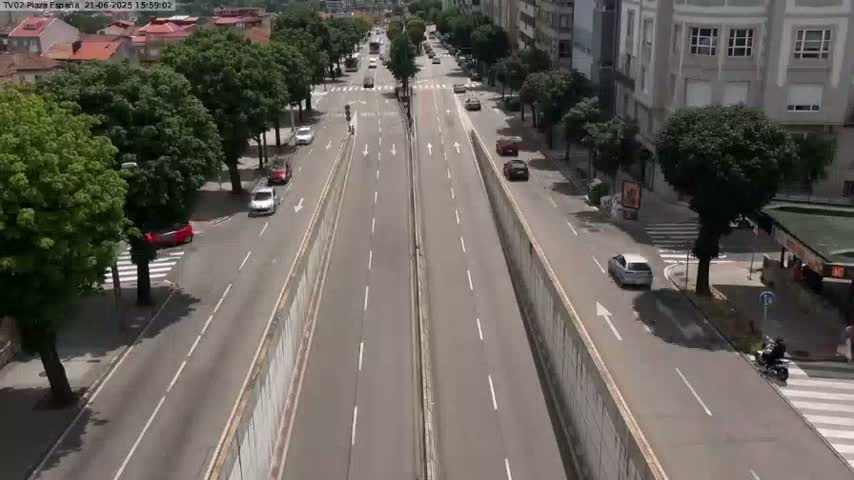 A sunny day reveals a multi-lane road cutting through a city, lined with trees and buildings, with several cars traveling in both directions.