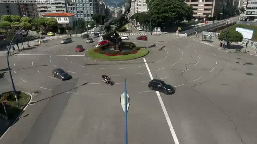 Several cars and motorcycles navigate a roundabout in a sunny city setting, centered around a large sculpture.