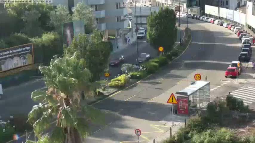 A sunny day overlooking a roadway with parked cars and a few vehicles in motion, near a building and lush greenery.