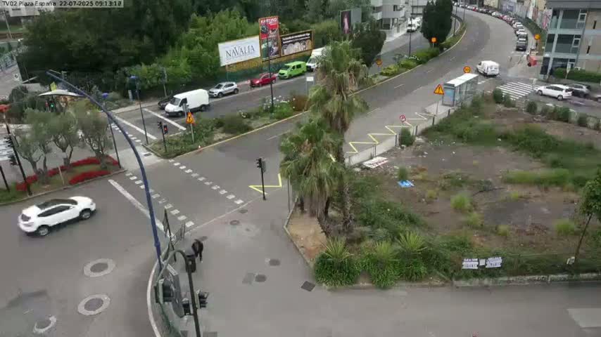 A high-angle view of a city intersection with several vehicles, palm trees, and an empty lot, under mostly cloudy skies.
